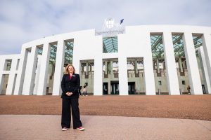 A woman with blonde hair wearing a black suit and red shoes, standing in front of Parliament House in Canberra.