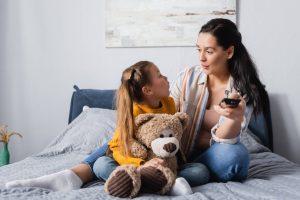 A girl is talking to her mother on a bed. The girl is wearing a yellow jumper and pigtails. The mother is holding a television remote and has long, black hair.