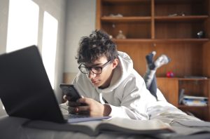 Young boy lying on a bed using computer and smartphone.
