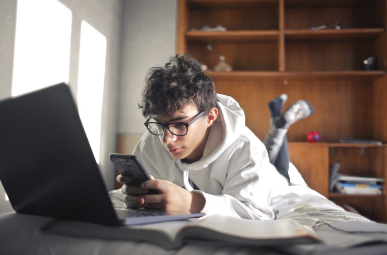 Young boy lying on a bed using computer and smartphone.