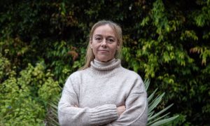 a woman with blonde hair wearing a cream jumper stands with arms crossed.