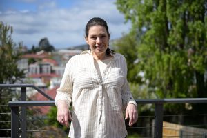 A woman is standing on a balcony, with her back and arms resting on a railing. In the background, you can see suburban Hobart.