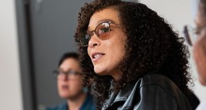 A woman with curly black hair wearing glasses and a leather jacket.