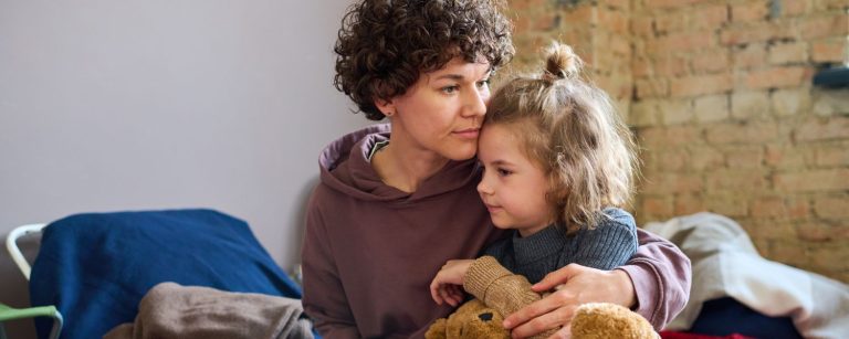 Young woman giving hug to her son who has a teddybear while both sitt on a bed.