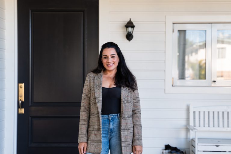 Maz Compton, a woman with long brown hair and a dark complexion, is standing in front of her house, which has a large black front door. Maz is wearing a patterned blazer, black top and blue jeans.