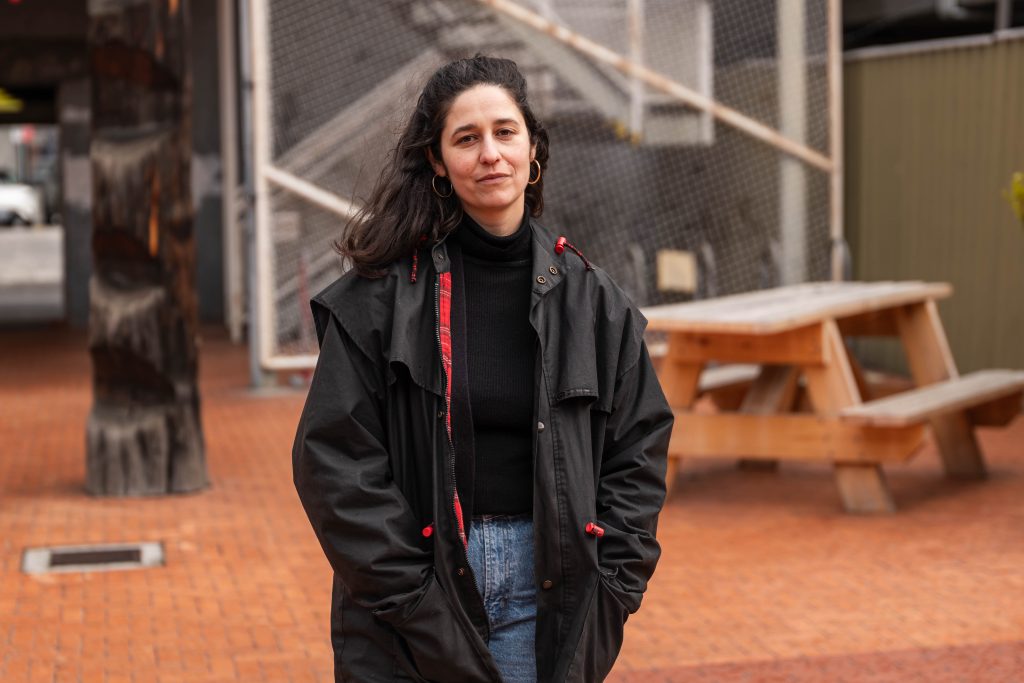 Natalya is pictured standing in front of a park bench on a tiled floor outdoors. Natalya is a woman with light skin, dark hair, and is wearing a large coat and jeans.