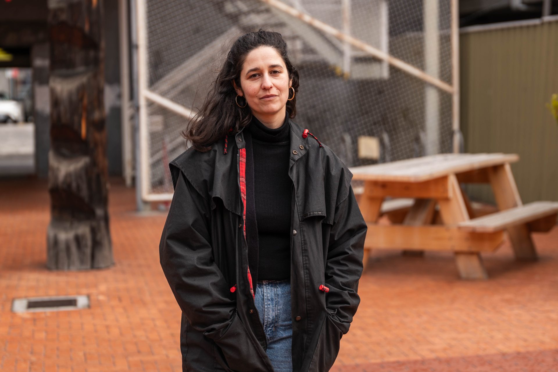 Natalya is pictured standing in front of a park bench on a tiled floor outdoors. Natalya is a woman with light skin, dark hair, and is wearing a large coat and jeans.