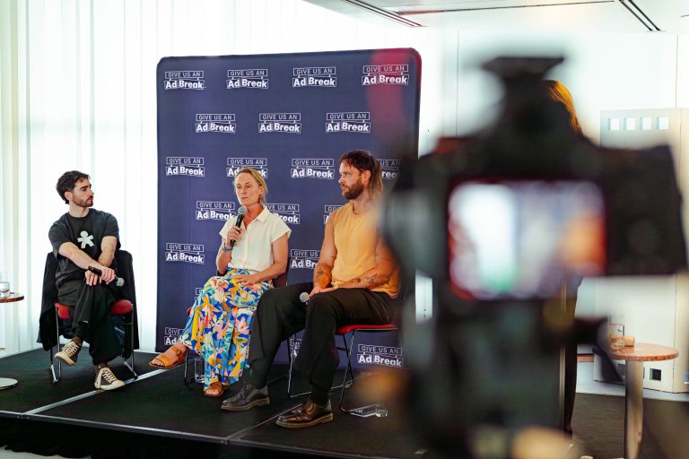 Kathryn Backholer, a woman with blonde hair and a multicoloured skirt, speaks from a microphone on a stage. A camera can be seen recording the discussion in the foreground.