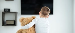 A child with blonde hair hugs a teddy bear while watching TV