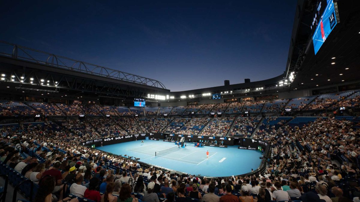 An open-air stadium with a blue tennis court in the centre, during the Australian Open. Two players are in the middle of a rally.